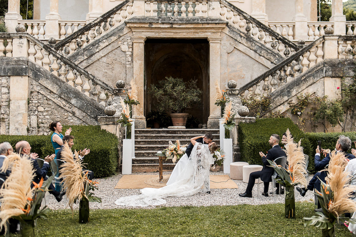 A bride and groom kiss at an outdoor wedding ceremony in front of a grand staircase, while guests seated on either side clap and watch.