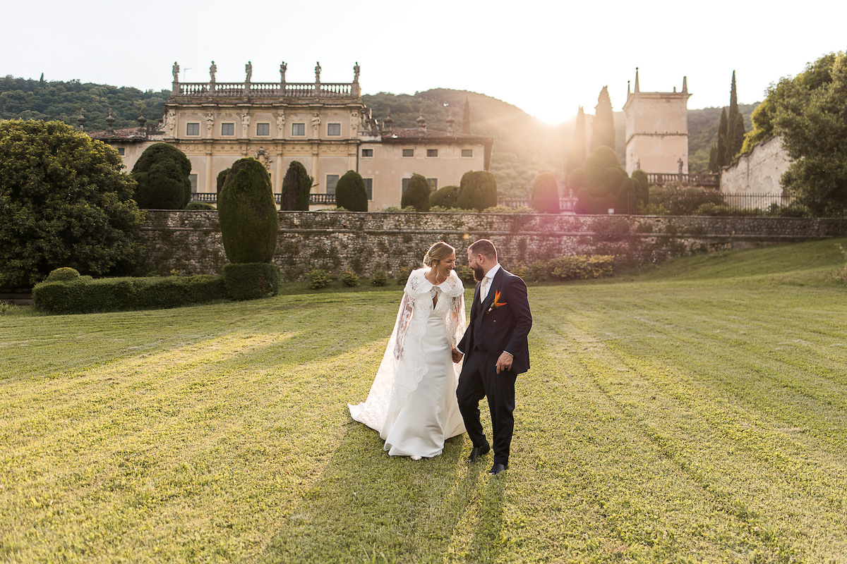 A bride and groom walk hand in hand across a sunlit lawn, with a historic building and trees in the background.