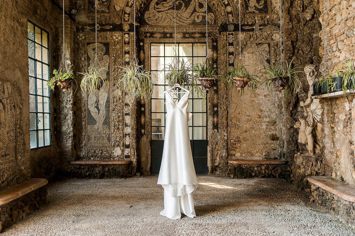 A white wedding dress hangs from the ceiling in the center of a rustic room with stone walls, mosaic details, and hanging plants.