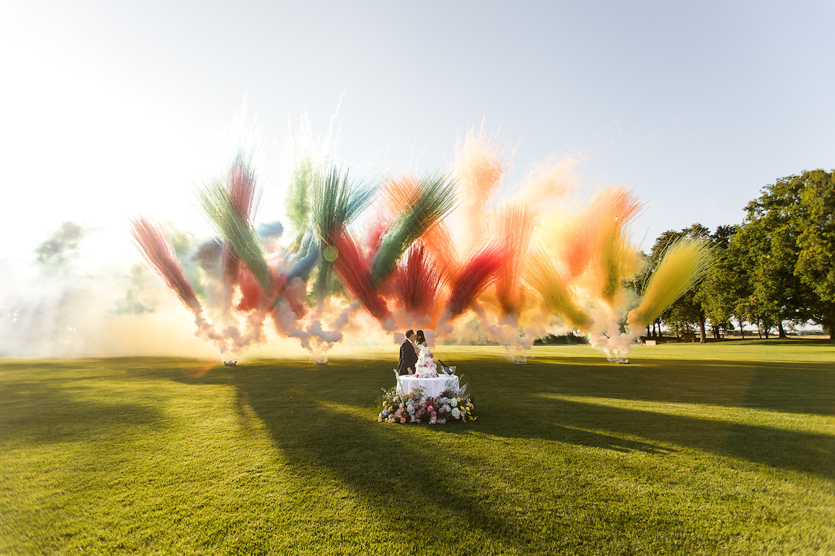 A couple stands by a decorated table on a grassy field as colorful smoke bursts into the air behind them under a clear sky.