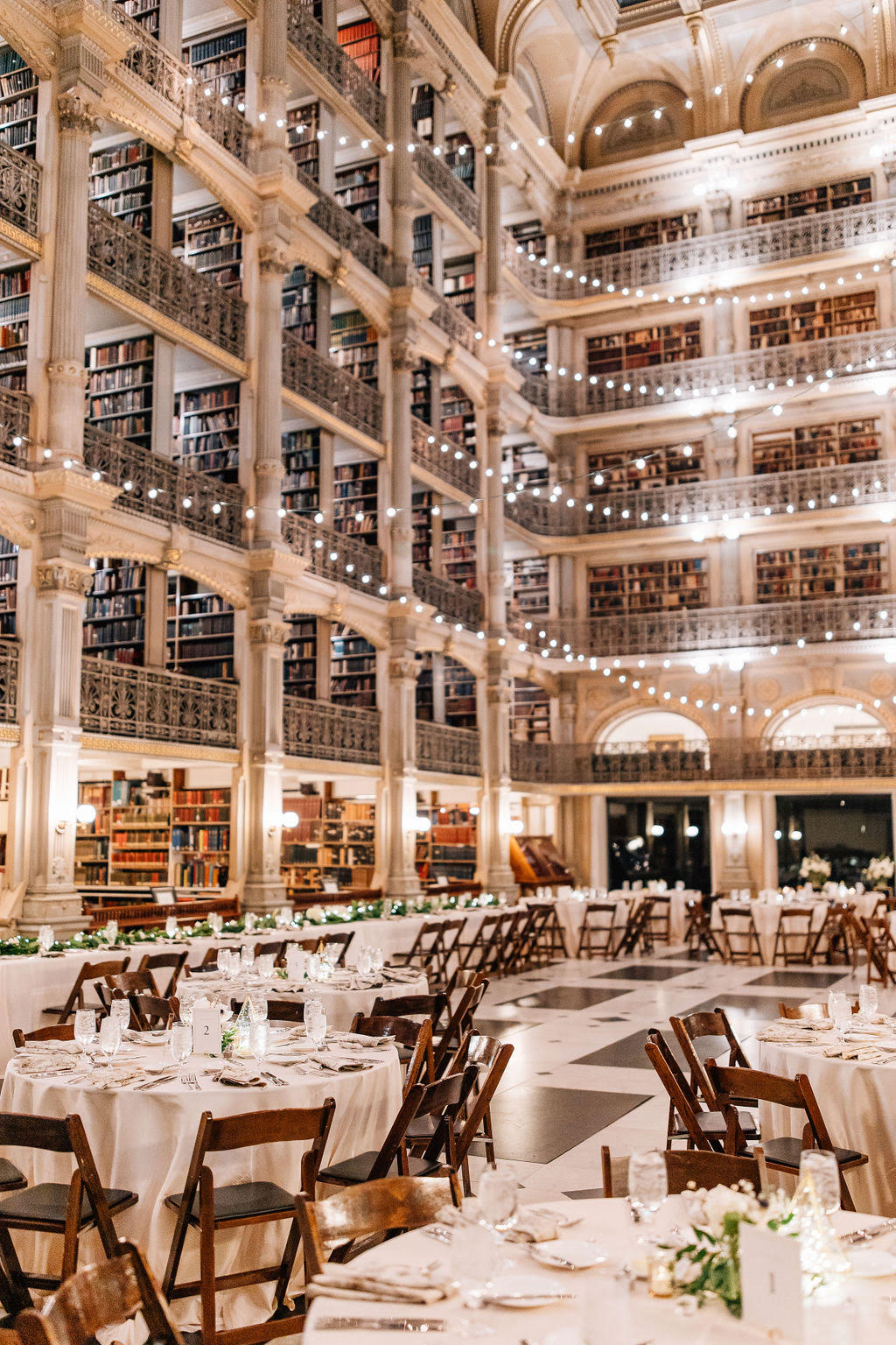 A grand library with multiple levels of bookshelves, decorated with string lights and set up for an event with round tables and wooden chairs.
