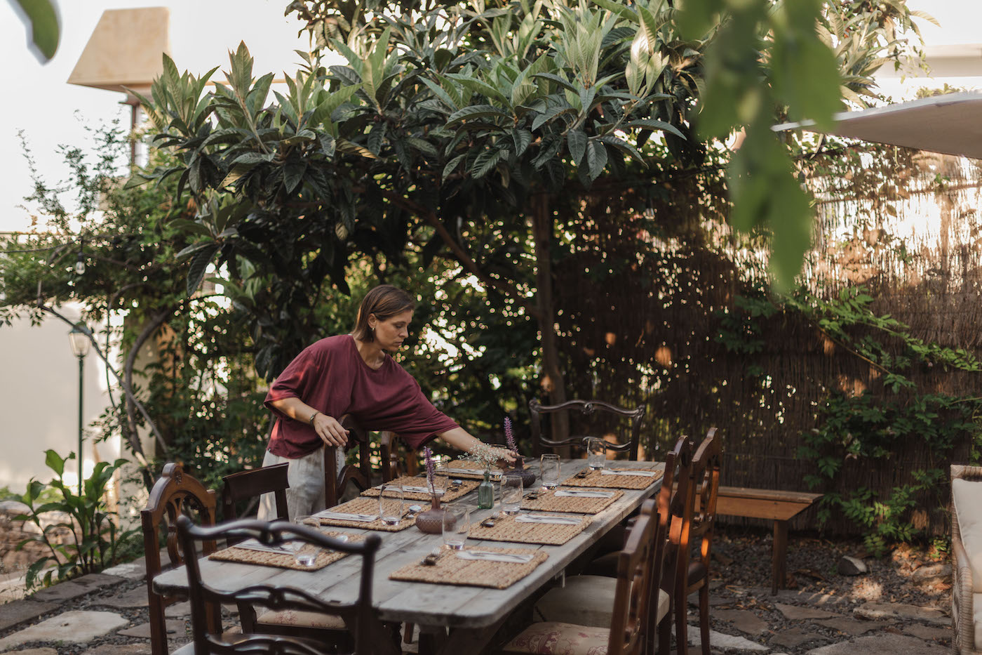 A person sets a wooden outdoor dining table with placemats and glasses in a garden surrounded by greenery.
