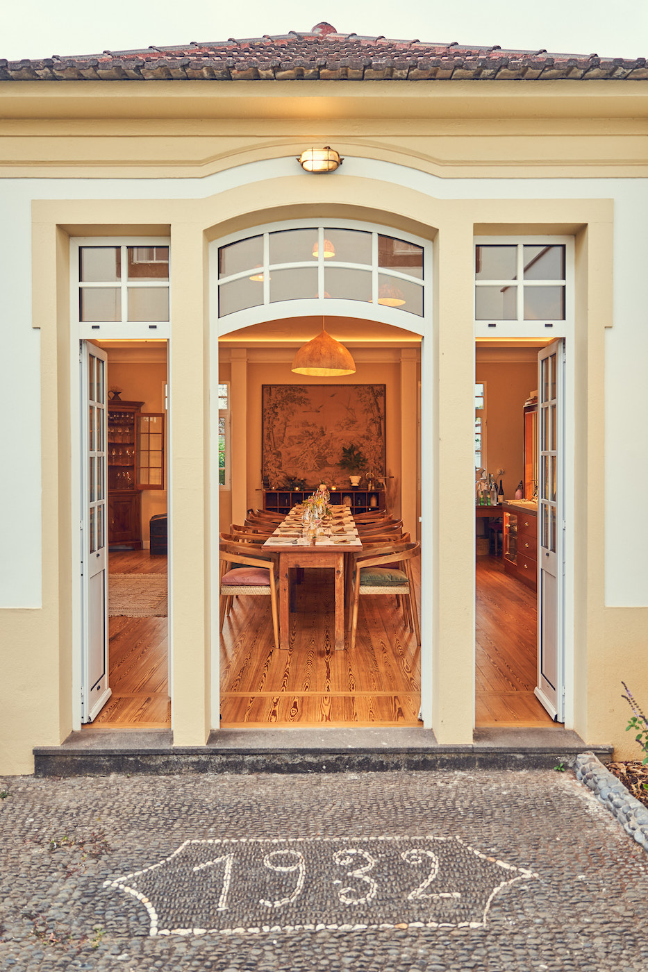 Open double doors reveal a warmly lit dining room set for a meal, with "1932" marked on the stone pathway at the entrance.