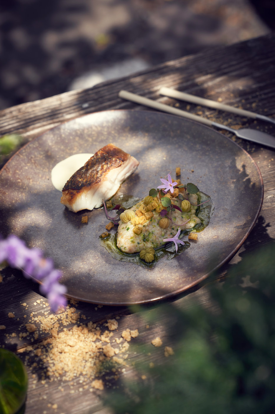 A plated dish of seared fish with a crisp skin, creamy sauce, green garnish, edible flowers, and crumbs, served on a round plate with cutlery on a rustic wooden table.
