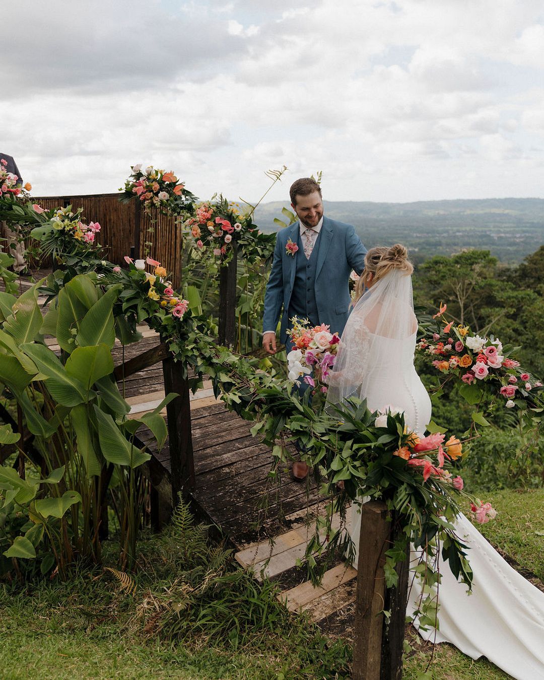 A bride and groom stand by a wooden staircase decorated with flowers and greenery, overlooking a scenic, hilly landscape under a cloudy sky.