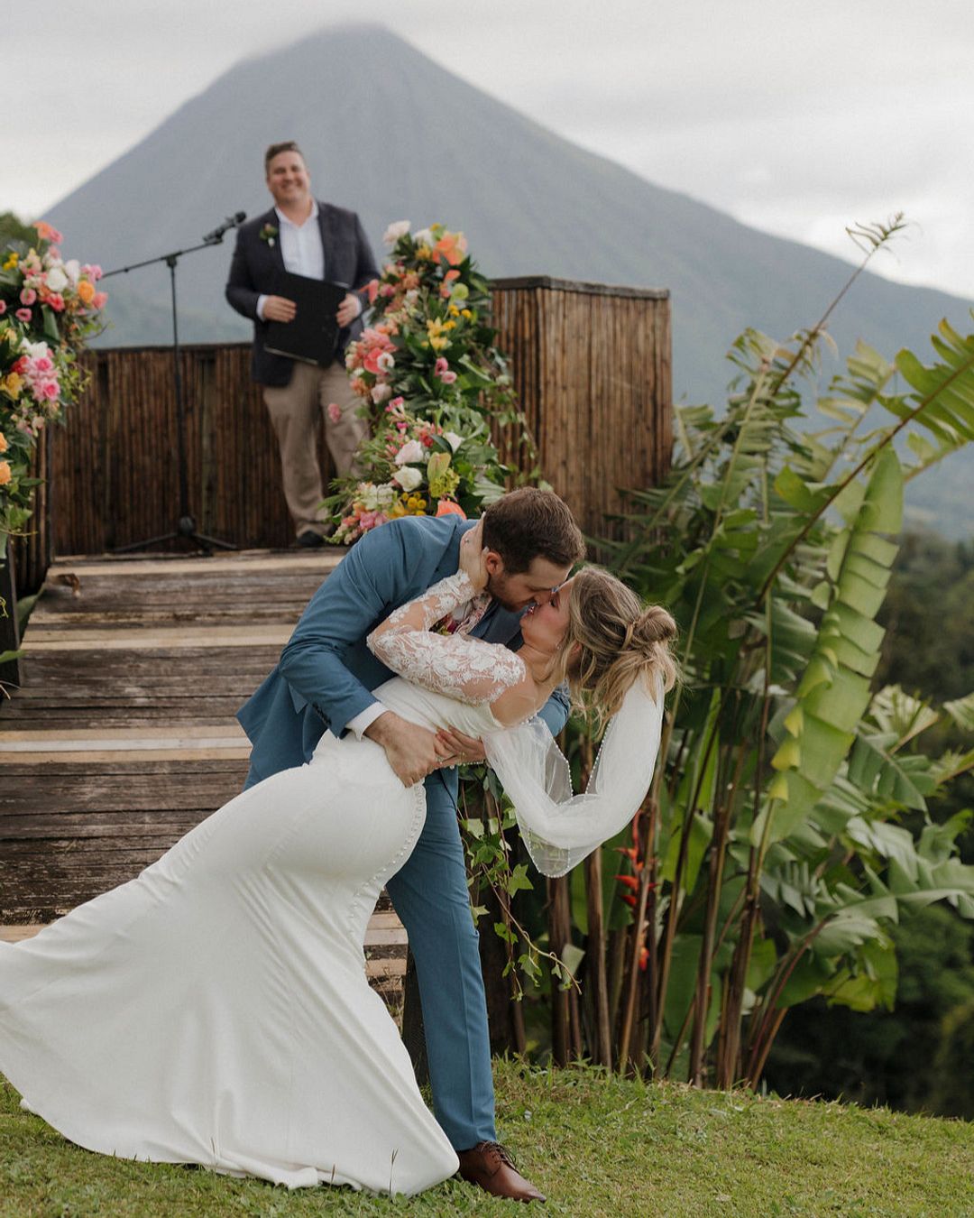 A bride and groom share a kiss outdoors in front of a wooden platform with flowers, as an officiant stands behind them, with a mountain in the background.