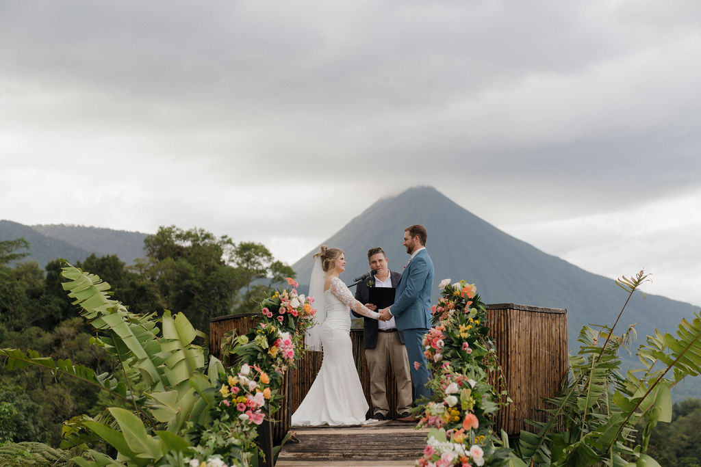 A bride and groom stand with an officiant on a flower-decorated platform outdoors, with a mountain visible in the background under a cloudy sky.