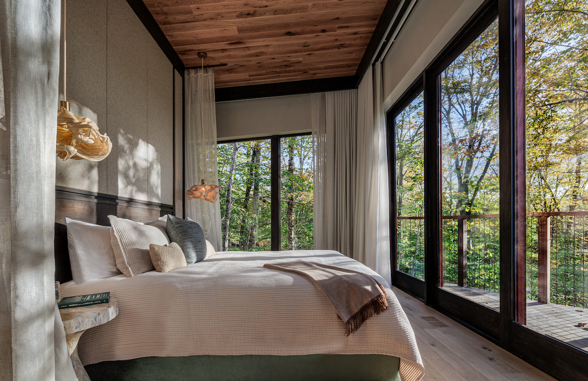 A modern bedroom with large windows overlooking a forest, featuring a wooden ceiling, neutral bedding, and natural light filling the space.