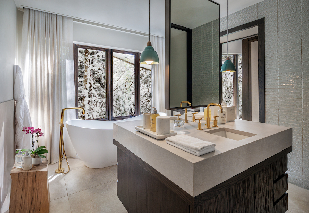 Modern bathroom with a freestanding bathtub by large windows, a double-sink vanity with gold fixtures, and hanging pendant lights.