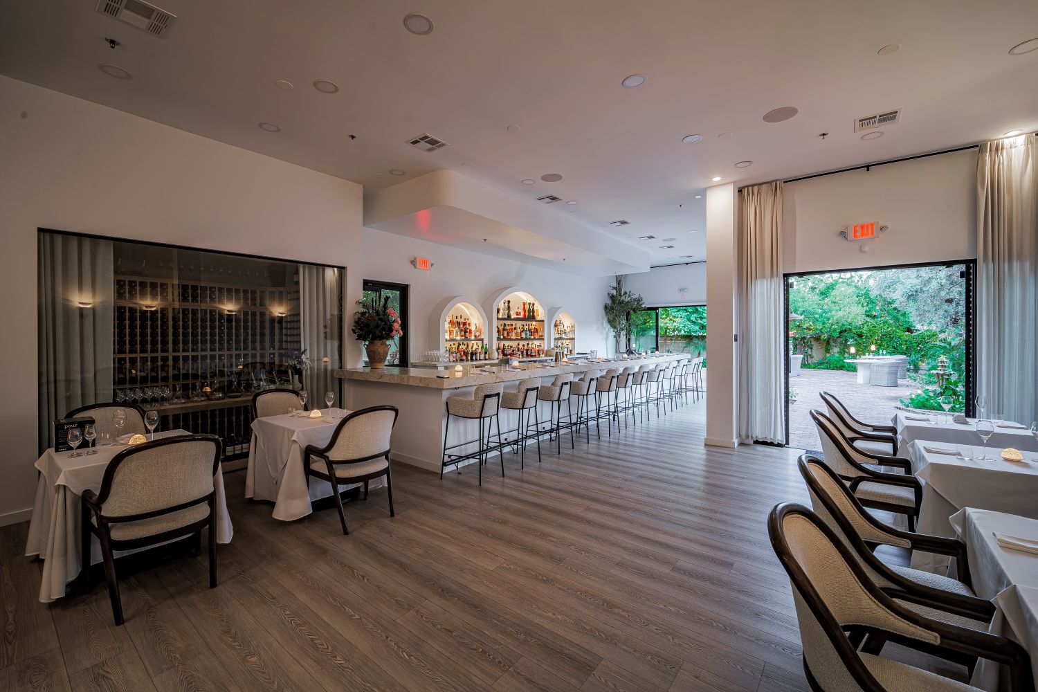 A modern restaurant interior with neatly arranged tables, a long bar with stools, light wooden floors, and large windows opening to a patio with greenery.