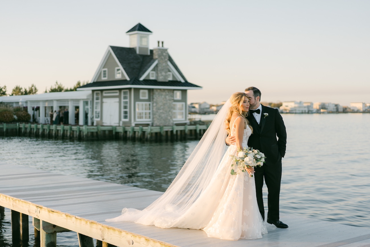 Bride and groom stand on a dock by the water, with the groom kissing the bride's forehead. A house on the shore is visible in the background.