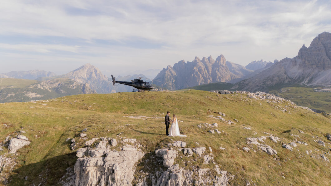 A bride and groom stand on a grassy mountaintop with a helicopter parked behind them and mountains in the background.