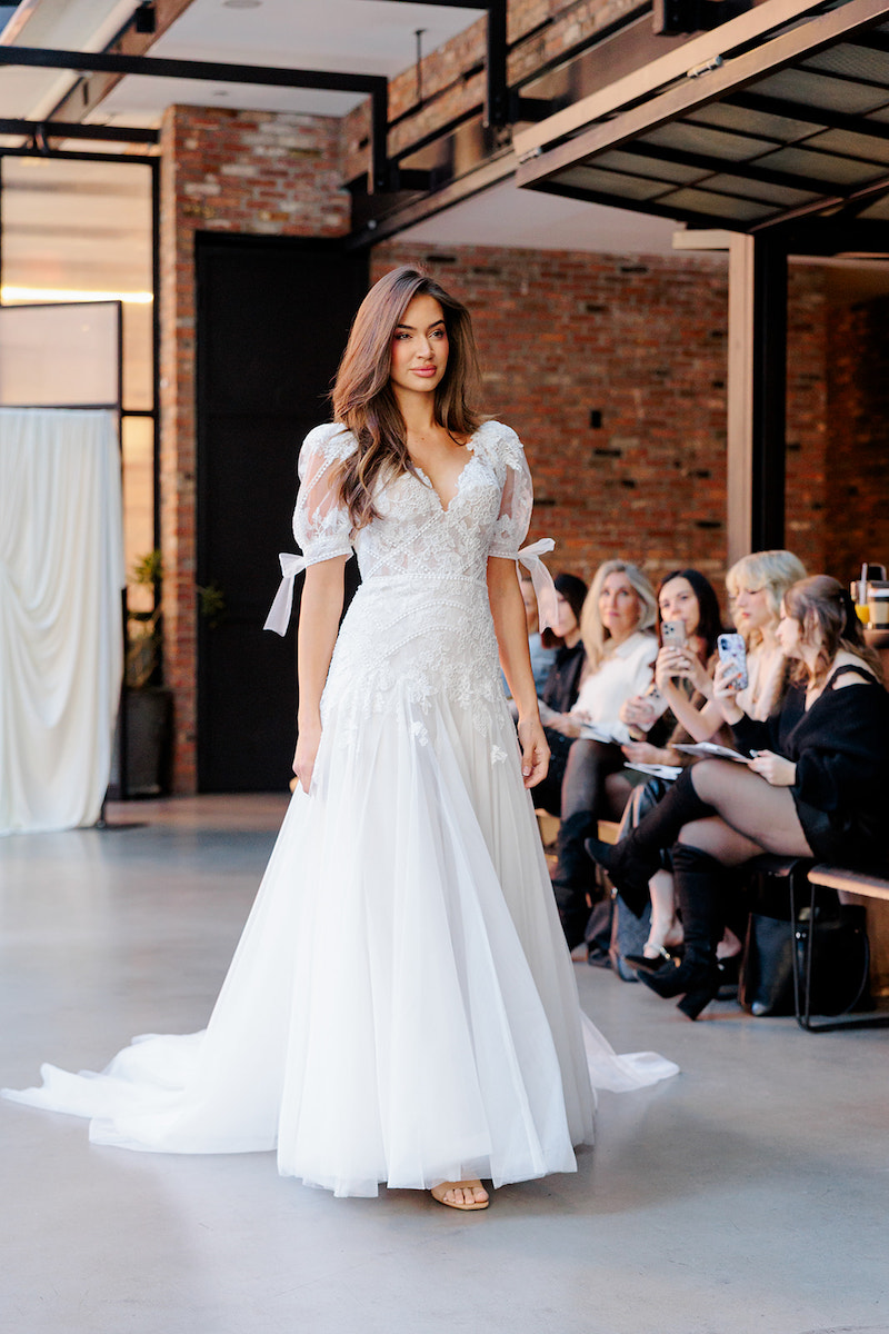 A model walks indoors wearing a white, floor-length wedding dress with lace details and puffed sleeves, while an audience seated on benches watches.