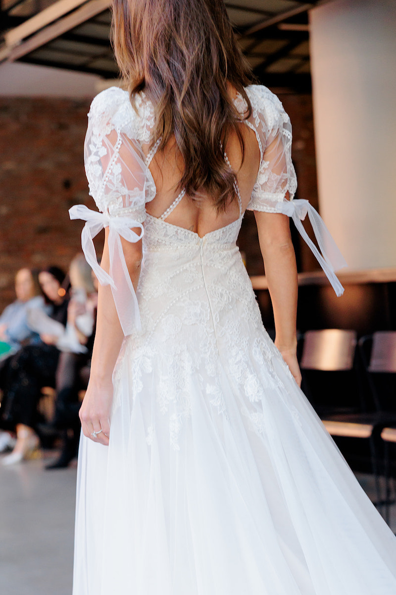 A woman in a white lace wedding dress with short puffed sleeves and ribbon ties is standing indoors, facing away from the camera.