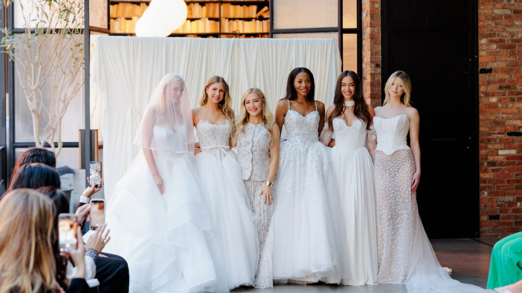Six women wearing white bridal gowns pose together indoors at what appears to be a fashion event, with seated audience members visible in the foreground.