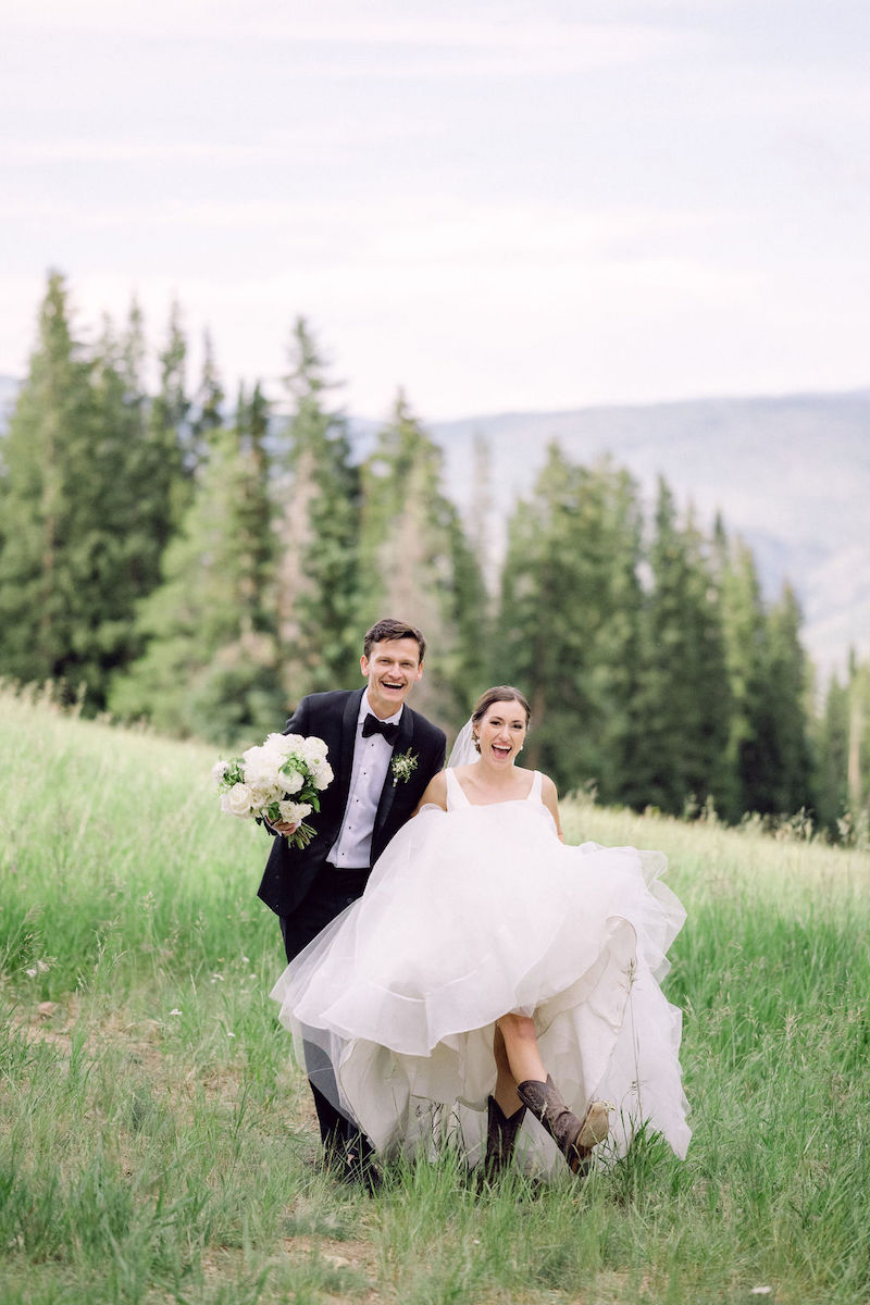 A bride in a white dress and boots is carried by a groom in a tuxedo as they walk through a grassy field with trees and mountains in the background.