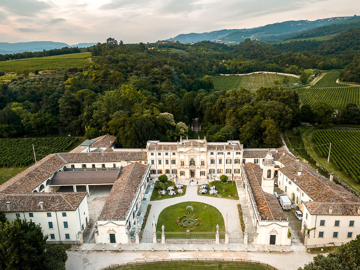 Aerial view of a large, historic villa with a central courtyard, surrounded by gardens, vineyards, and rolling hills.
