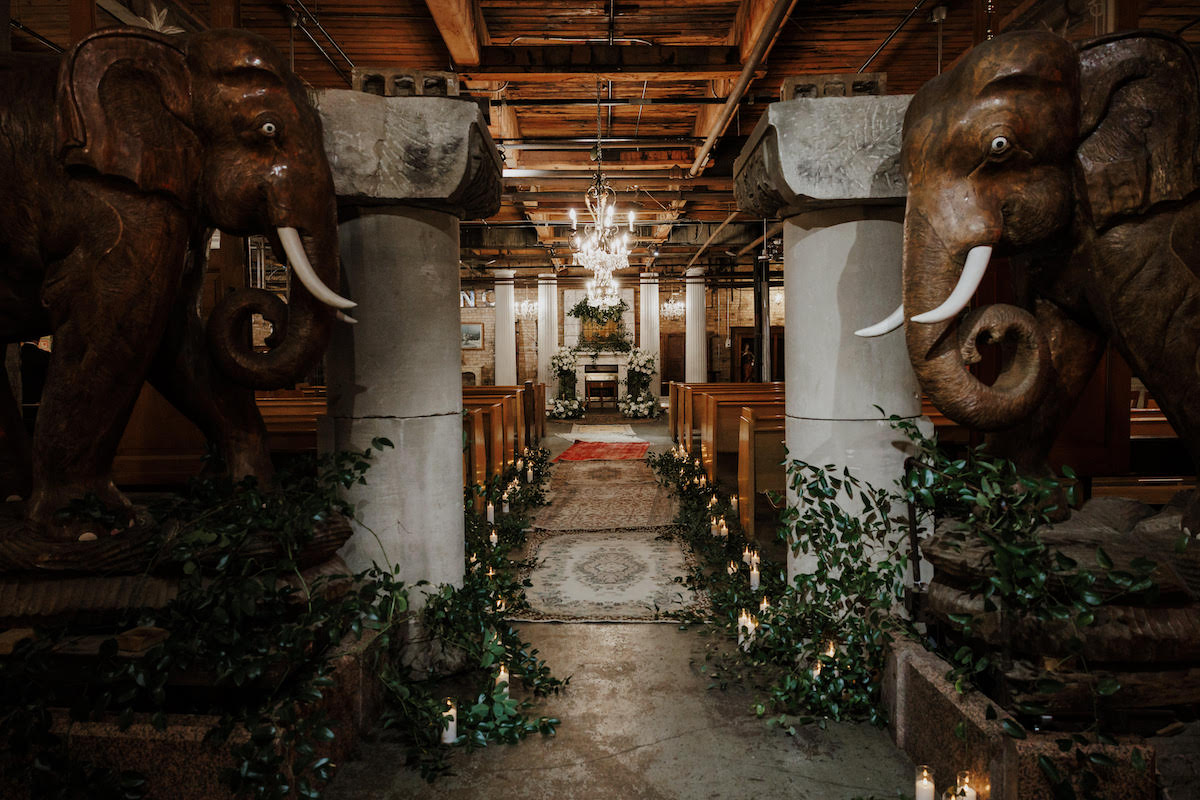 A grand indoor aisle flanked by large elephant statues, columns, greenery, and candles, leading to an altar under a chandelier.