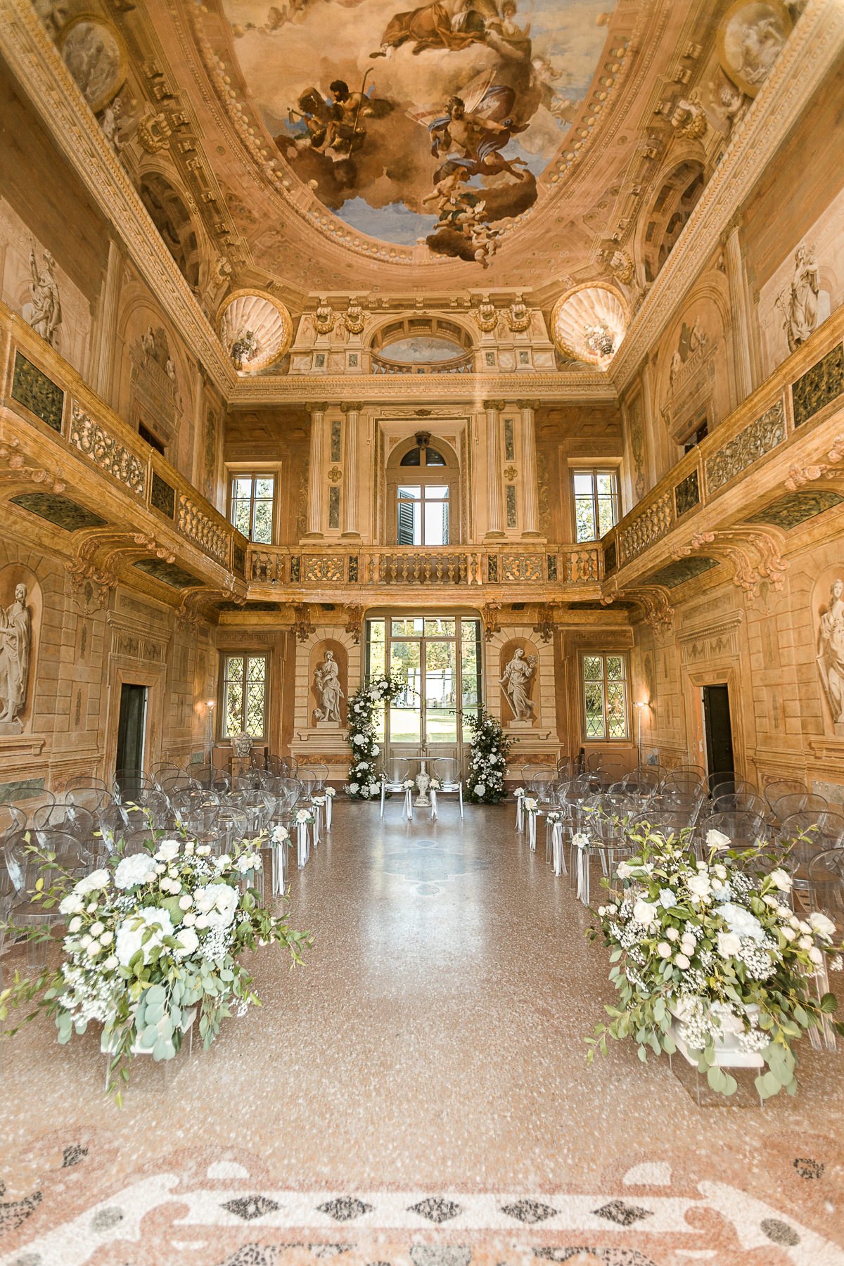 Lavishly decorated hall with ornate ceiling fresco, marble statues, large windows, and elegant floral arrangements set up for a formal event or ceremony.