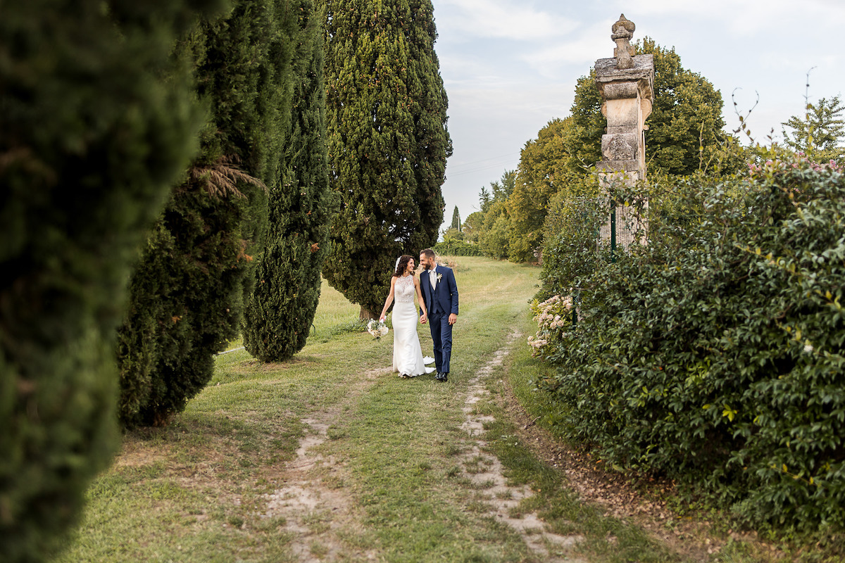 A bride and groom walk together on a grassy path lined with tall trees and greenery, with a stone monument visible on the right.