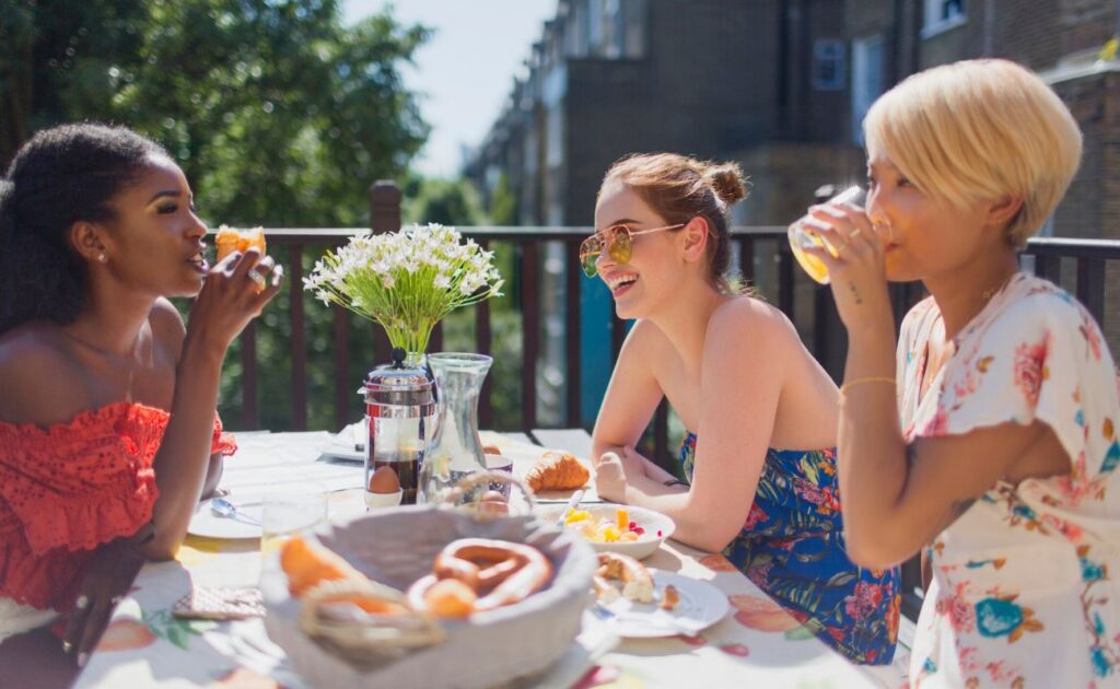 Three people sit at an outdoor table with food and drinks, talking and smiling. A vase of white flowers is in the center of the table.