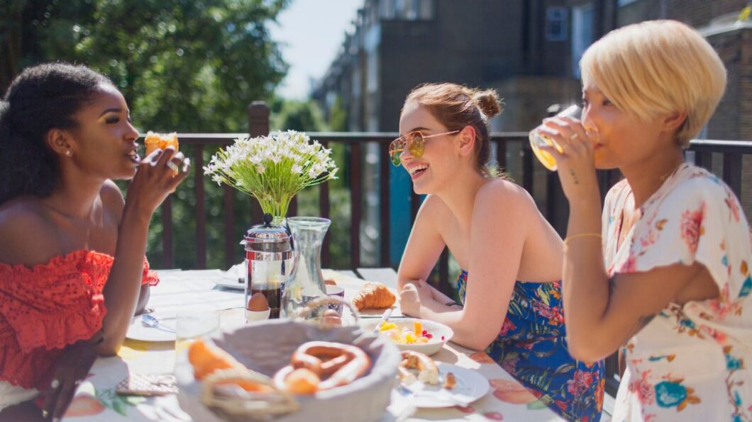 Three people sit at an outdoor table with food and drinks, talking and smiling. A vase of white flowers is in the center of the table.