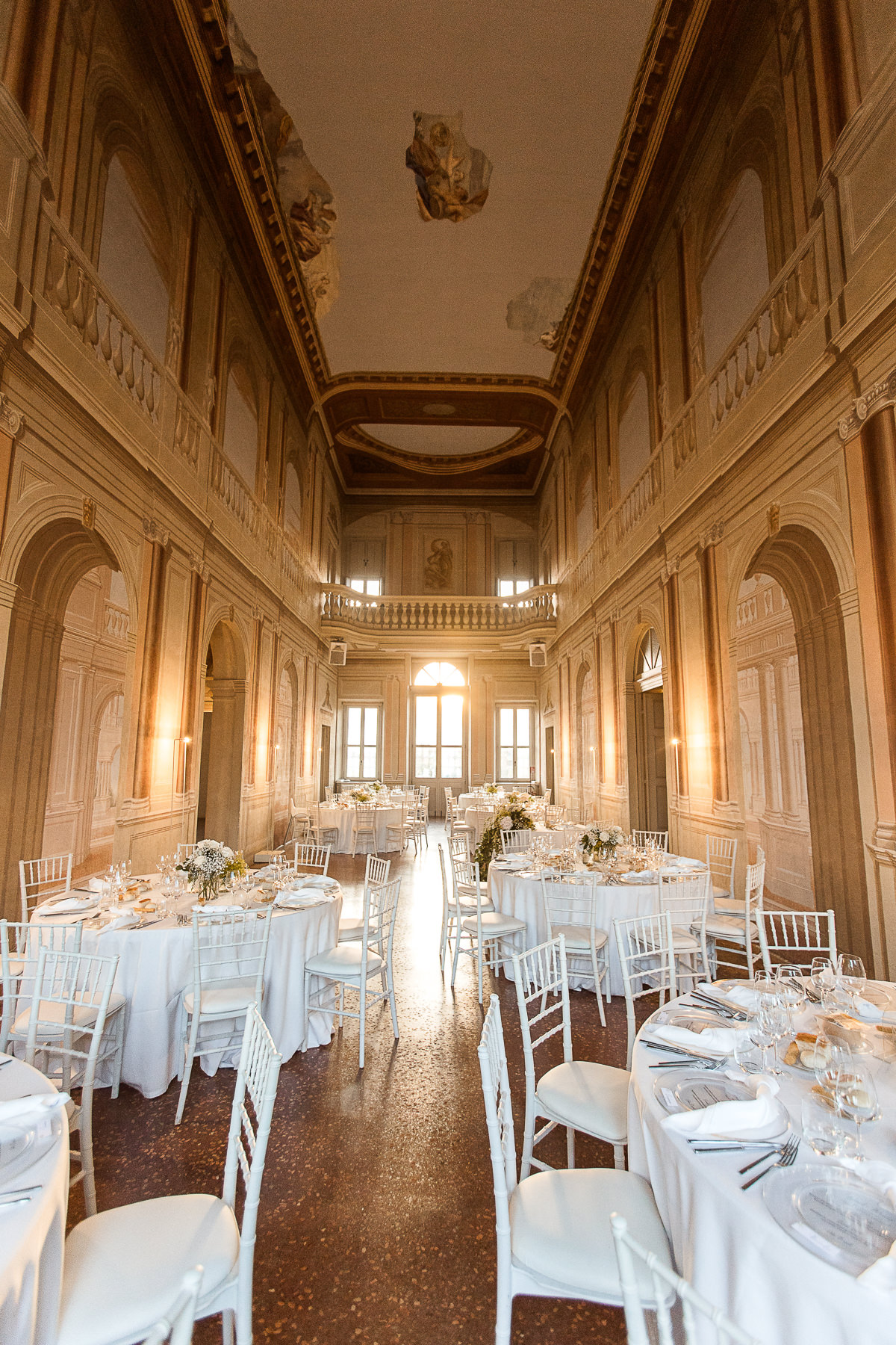 Elegant banquet hall with high ceilings, ornate walls, and multiple round tables set with white linens, plates, and glassware, ready for a formal event or reception.