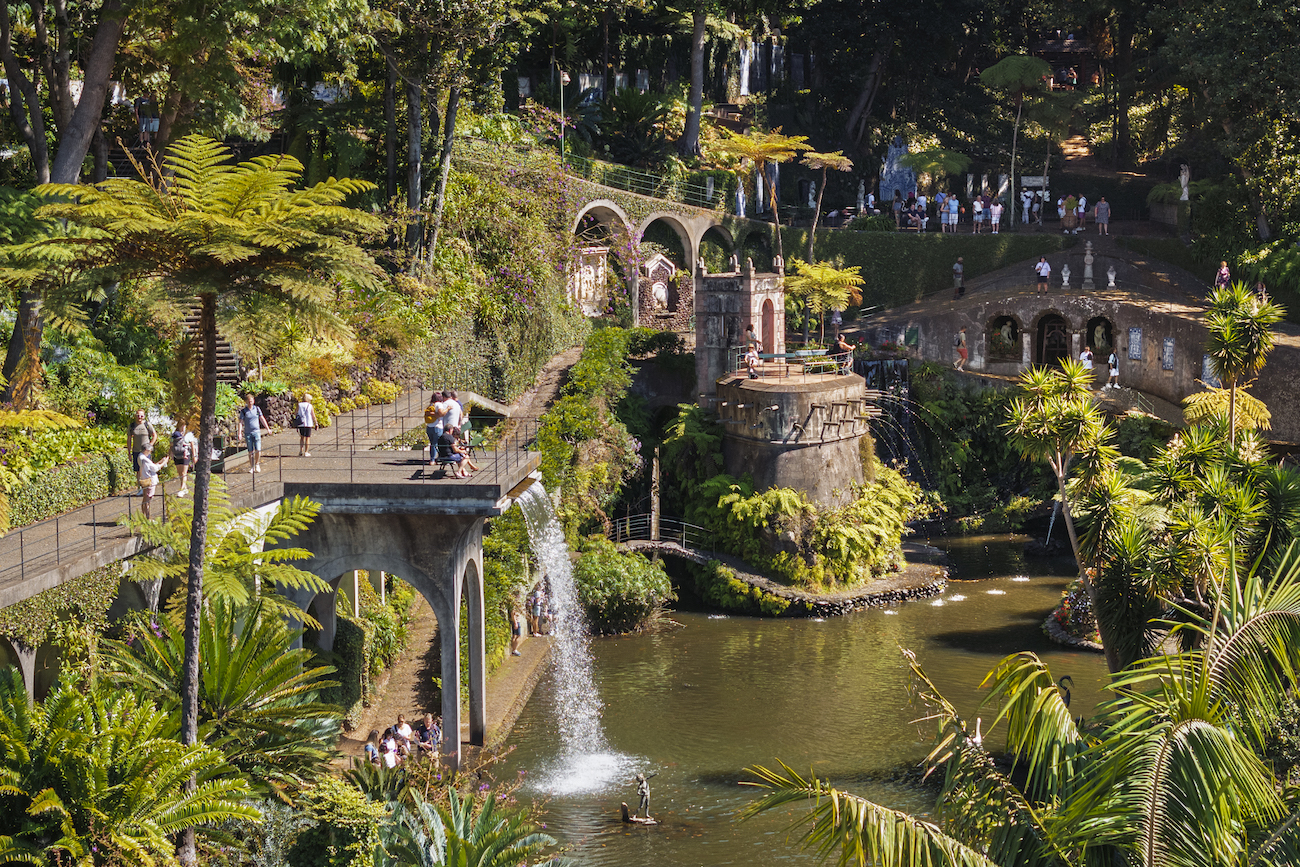 Lush botanical garden with stone bridges, a small waterfall, a pond, and visitors walking along pathways amid dense greenery and sculptures.
