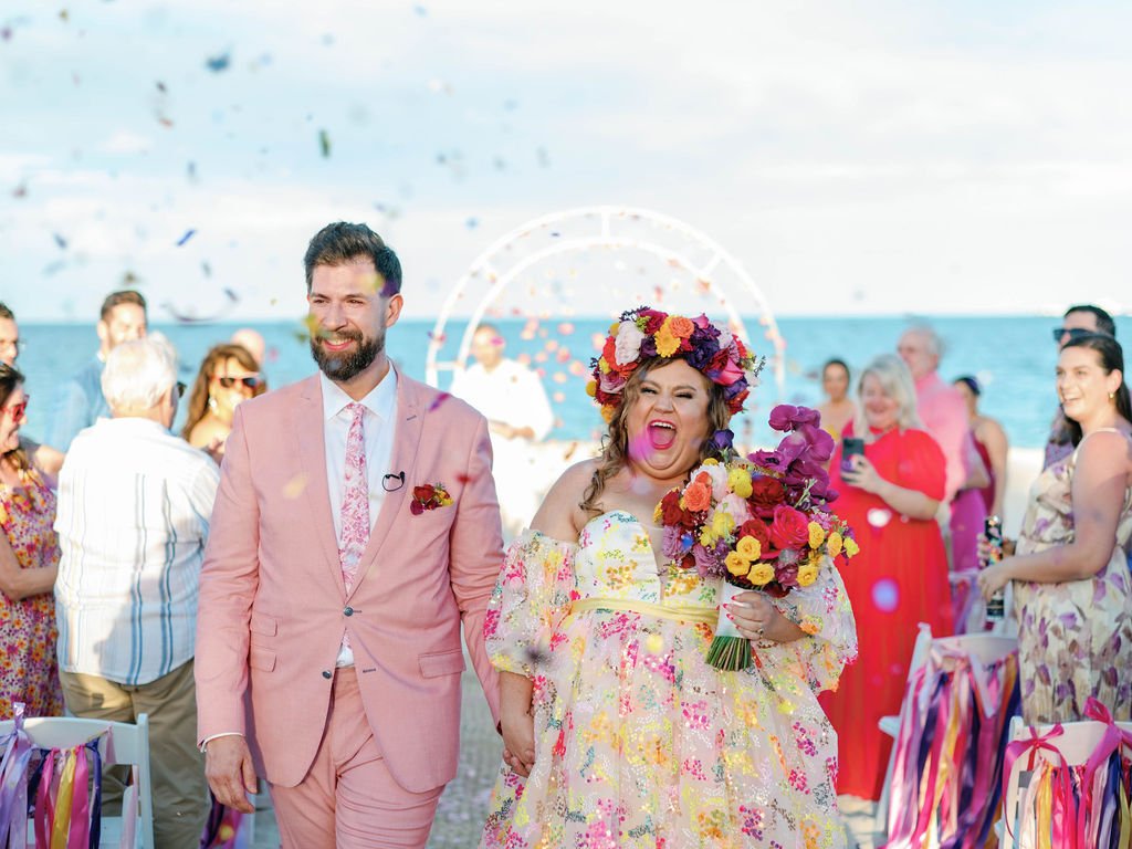 A couple walks down the aisle at an outdoor wedding by the sea, smiling and holding hands, with confetti in the air and guests in the background.