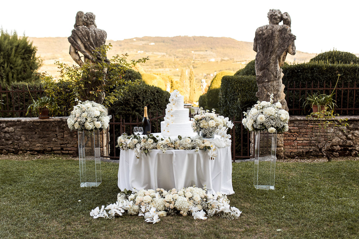 Outdoor wedding cake table with white floral arrangements and champagne, set between two stone statues in a garden.