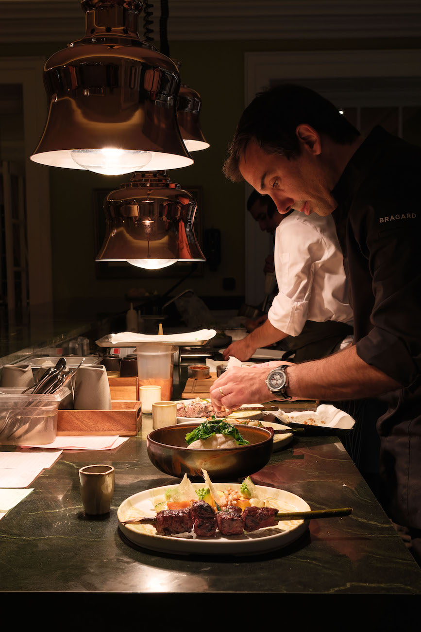 A chef is plating dishes under warm lights in a restaurant kitchen, with various ingredients and utensils arranged on the counter.