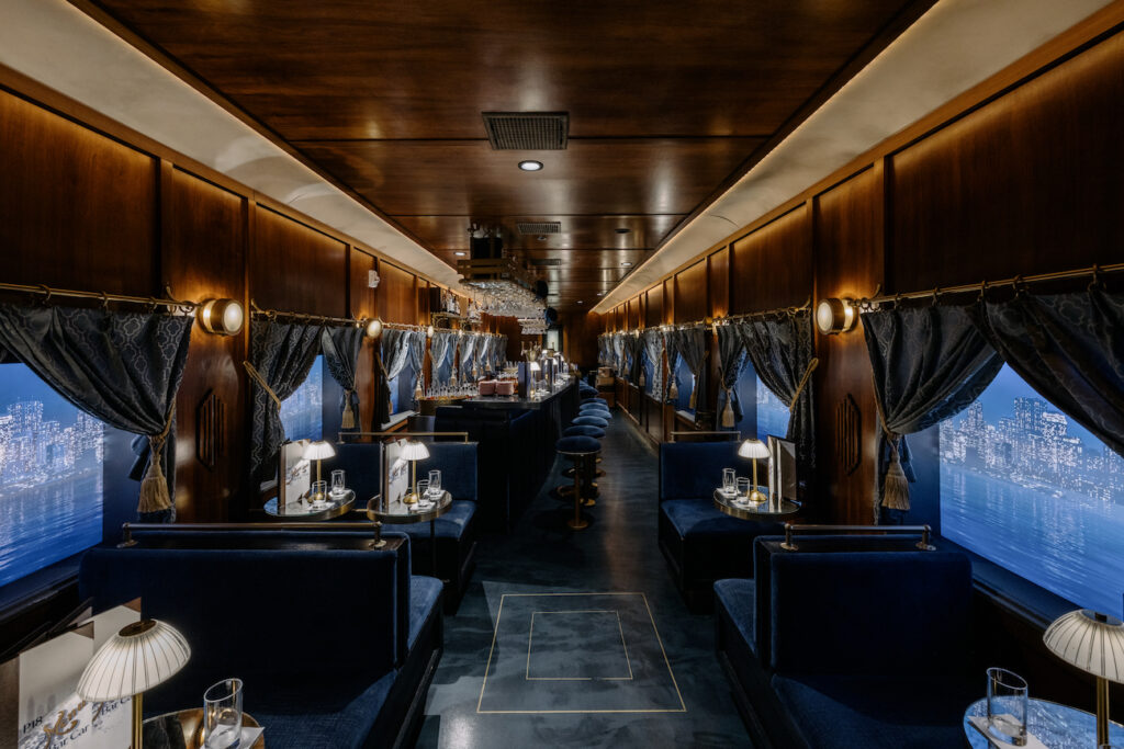 Luxurious train dining car with dark wood paneling, blue upholstered seats, small elegant lamps, and large windows with cityscape views.