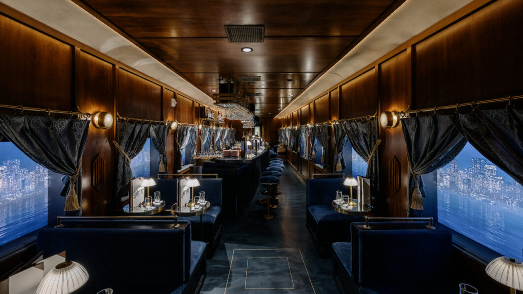 Luxurious train dining car with dark wood paneling, blue upholstered seats, small elegant lamps, and large windows with cityscape views.