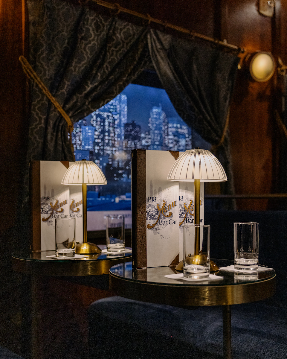 A small round table with two empty glasses and a lamp, in front of a window with city buildings visible at night. Dark curtains frame the window.