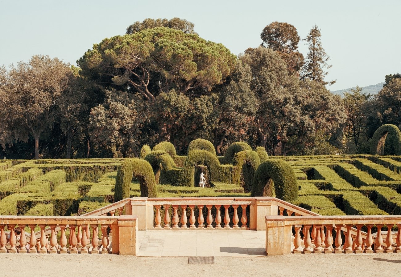 A manicured hedge maze with arched topiaries, viewed from behind a stone balustrade, with tall trees in the background.