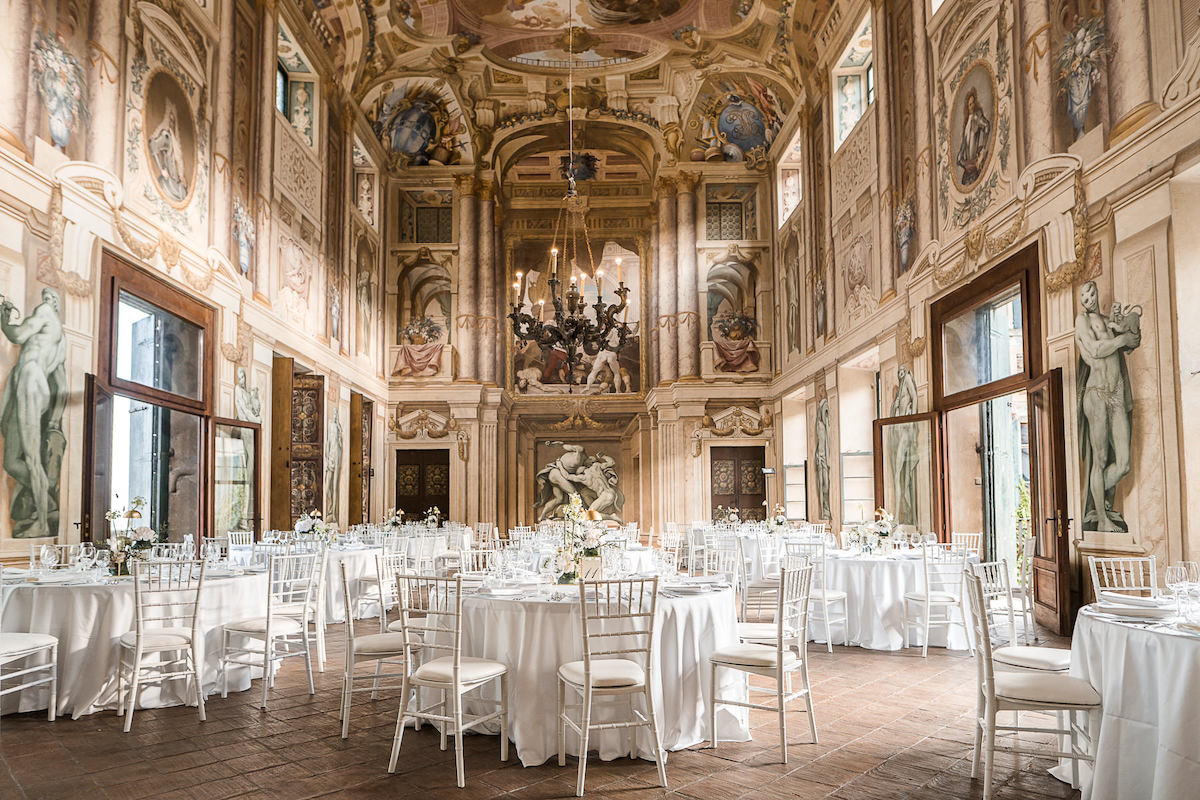 Elegant banquet hall with high ceilings, ornate wall and ceiling frescoes, round tables with white linens, and white chairs arranged for an event.