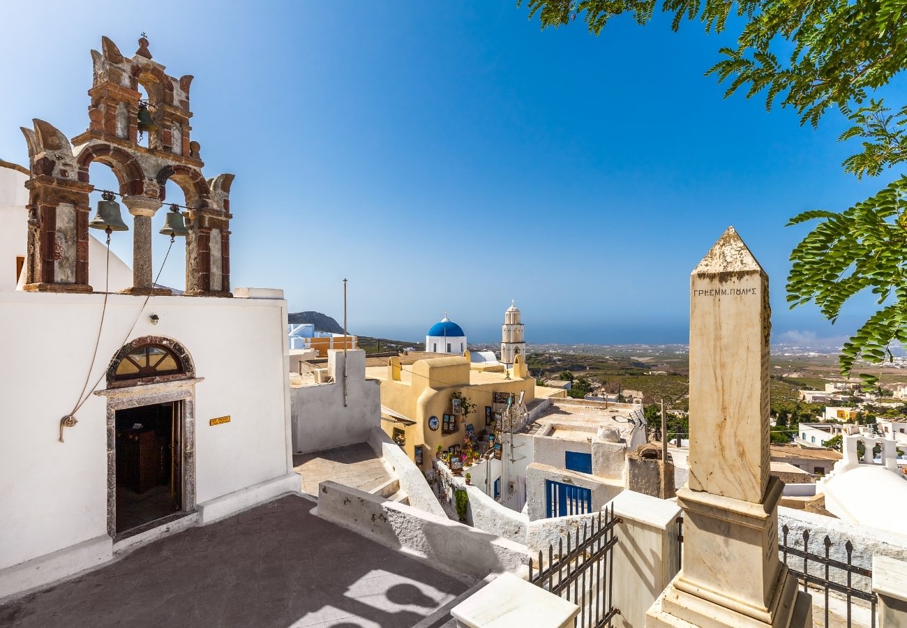 Stone bell tower and white buildings with blue domes overlook the sea under a clear sky in a Greek coastal village.