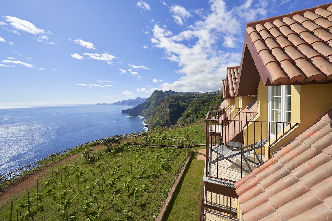 A coastal house with tiled roofs and balconies overlooks green vineyards, cliffs, and the ocean under a partly cloudy sky.
