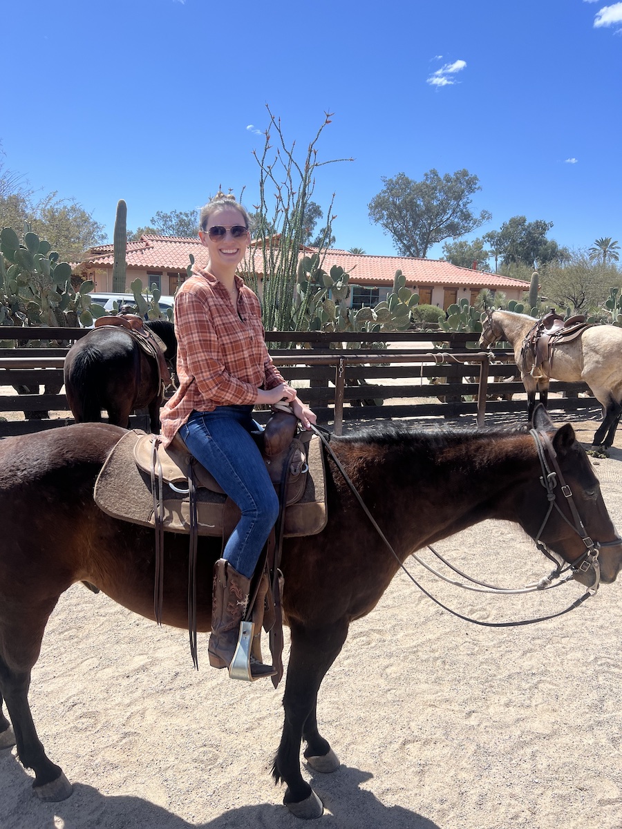 A woman wearing sunglasses, jeans, boots, and a plaid shirt sits on a brown horse in a sandy corral with cacti and trees in the background.