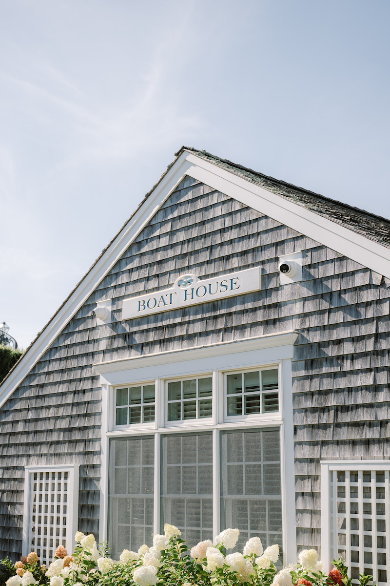 A gray shingled building with a sign reading "Boat House" above white-trimmed windows, set against a clear blue sky and surrounded by white flowers.