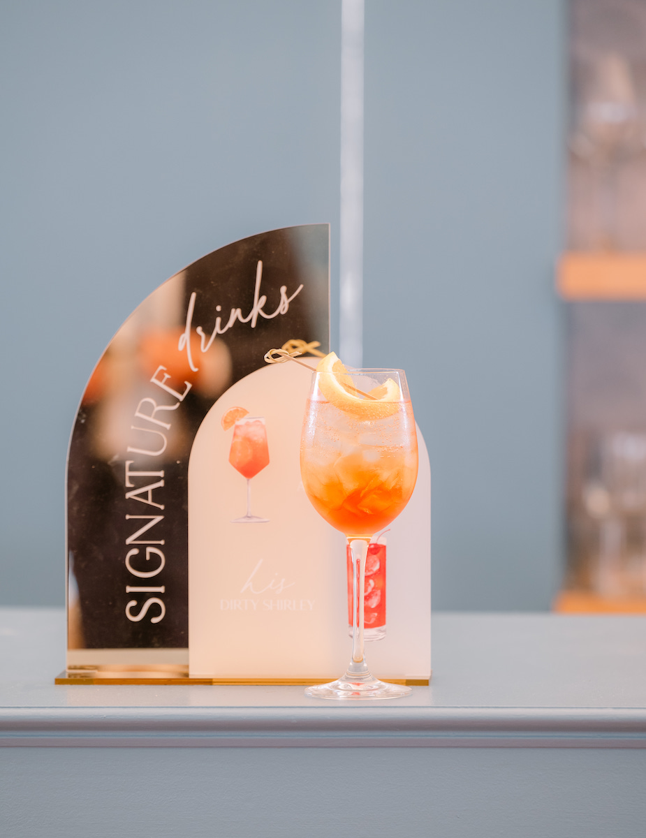 A glass of orange cocktail with ice and an orange twist sits on a counter next to a "Signature Drinks" sign.