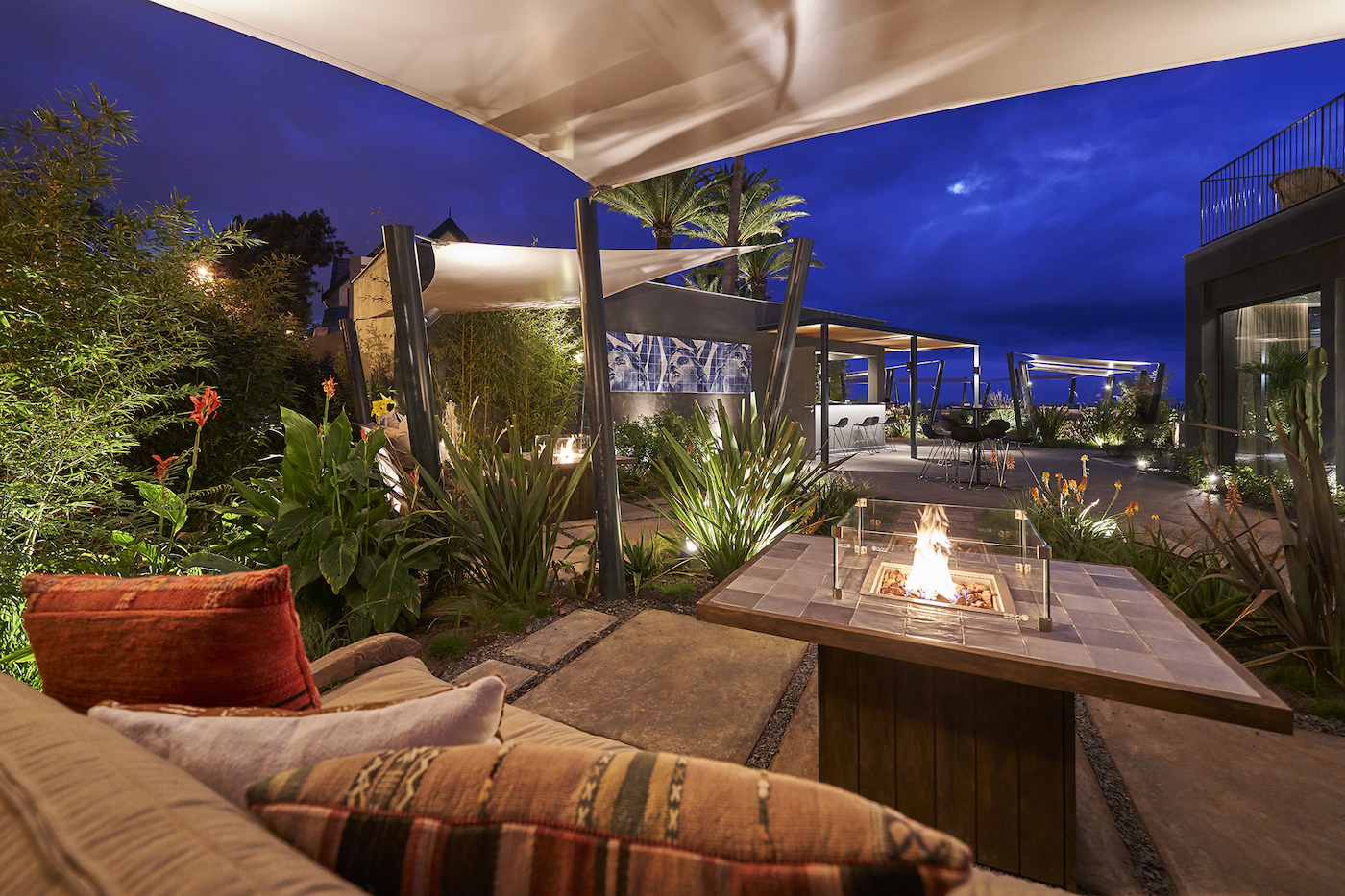 Outdoor patio at night with a modern fire pit, cushioned seating, lush plants, and canopy structures under a cloudy sky.