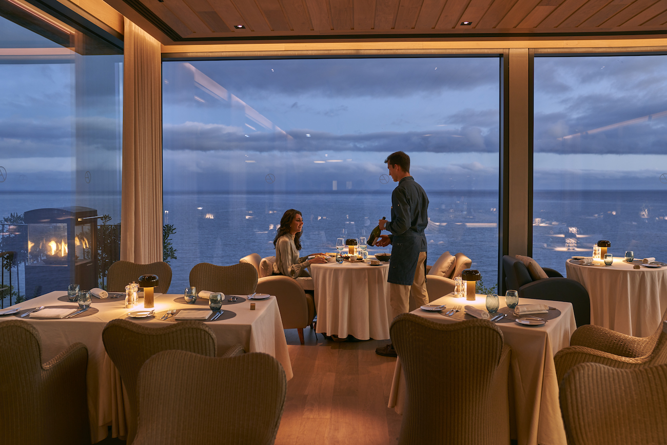 A server pours wine for a woman seated at a table in an upscale restaurant with large windows overlooking the ocean at dusk.
