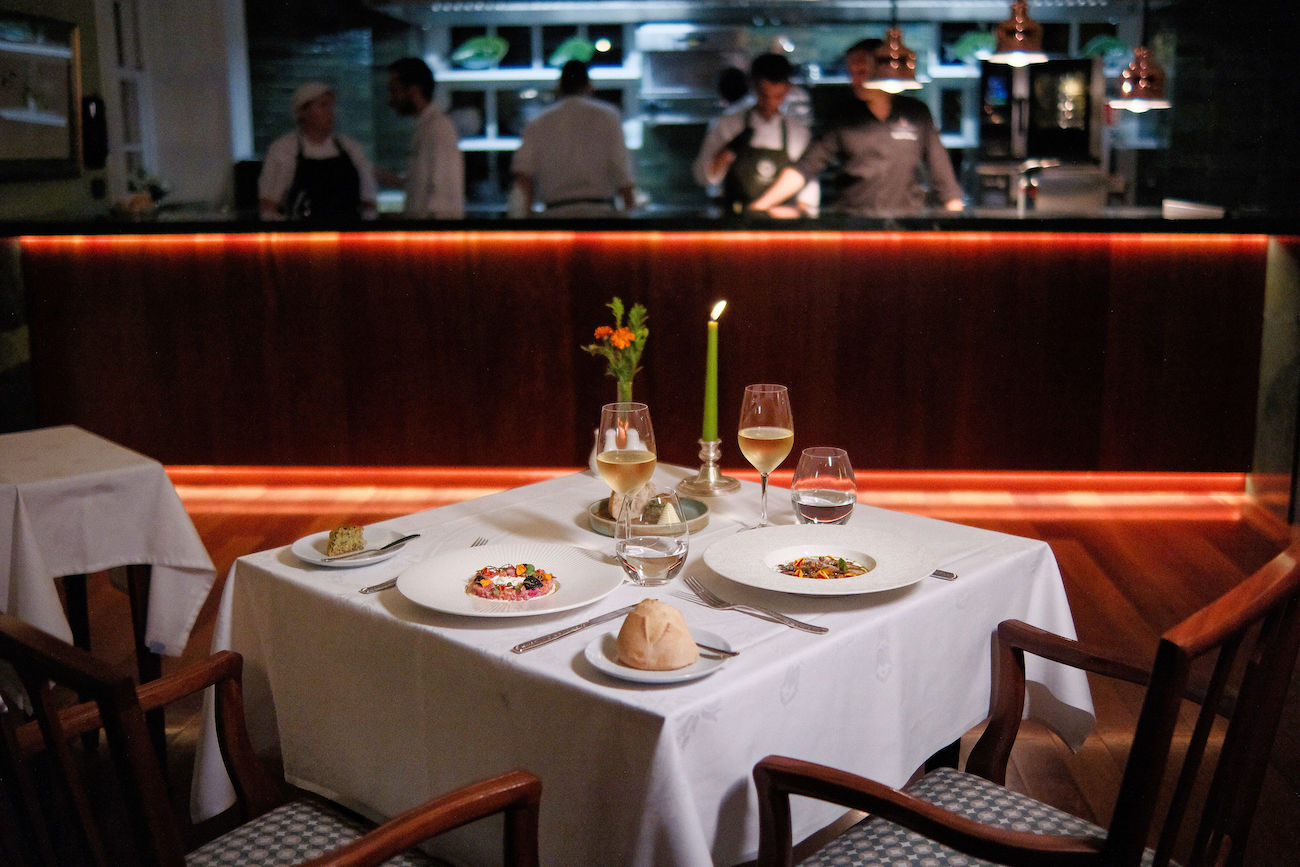 A restaurant table set for two with plated dishes, drinks, and a lit candle; chefs work in an open kitchen in the background.