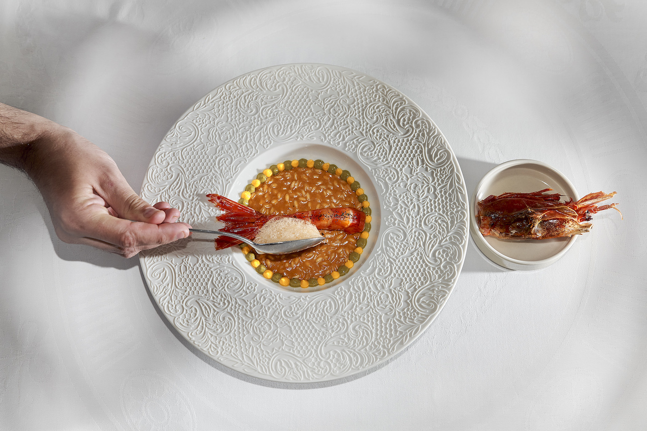 A hand places a spoon with foam over a dish of risotto topped with shrimp, with another shrimp served in a small bowl on the side.