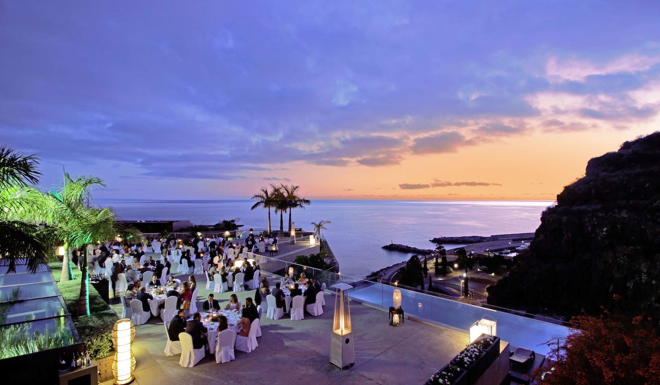 Outdoor evening event with guests seated at round tables near a pool, overlooking the ocean at sunset, with palm trees and mountains in the background.