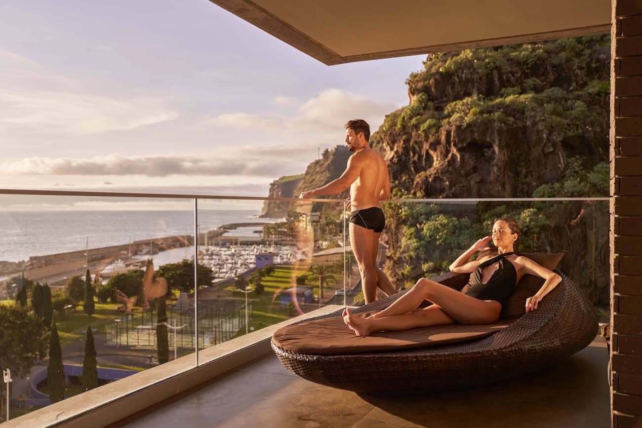 A man stands at a balcony railing overlooking the ocean while a woman lounges on a round outdoor daybed on a terrace with coastal views.