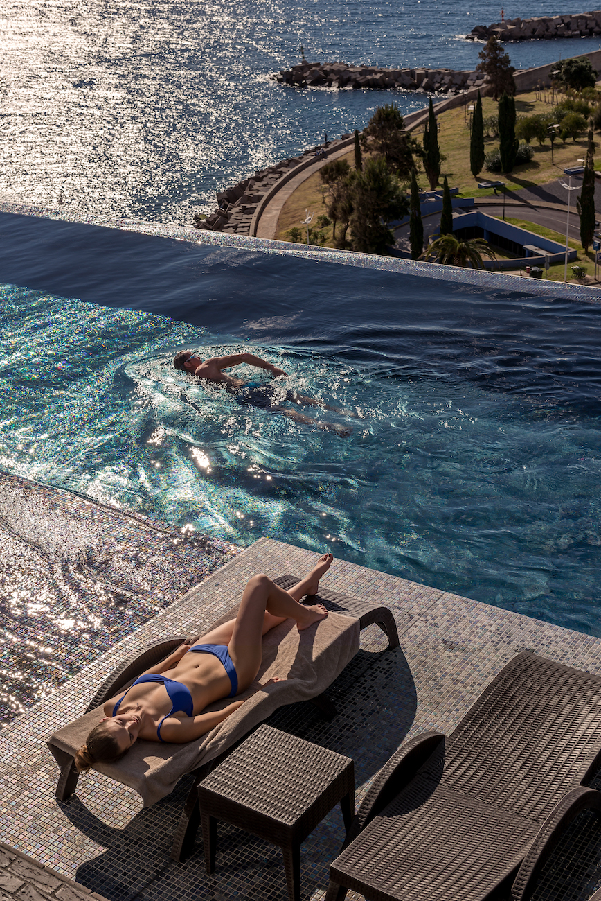A woman in a blue swimsuit lounges on a pool chair while a person swims in an infinity pool overlooking a coastal landscape and ocean.