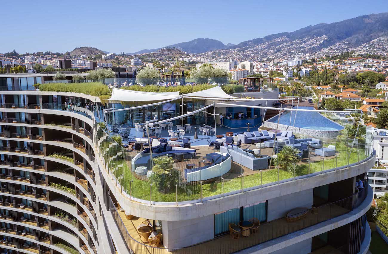 A rooftop lounge with seating areas, greenery, and a small pool overlooks a cityscape with hills and mountains in the background.