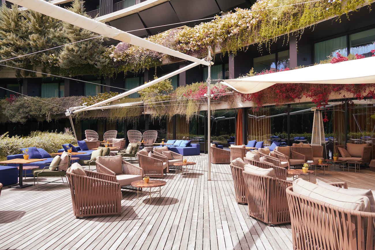 Outdoor hotel lounge area with wicker chairs, tables, and sofas on a wooden deck, shaded by awnings and surrounded by greenery and flowering plants.